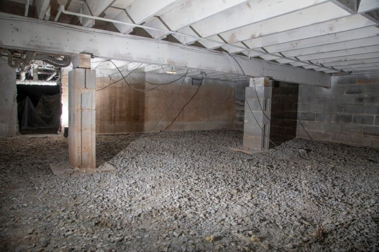 A dimly lit, unfinished basement with exposed beams and cinder block walls. The floor is gravel, wires hang from above, and pillars support the ceiling. Crawl Space Drainage Systems