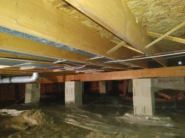 Wooden beams and cinder block piers support a house in a crawl space. Insulation and pipes are visible, creating an industrial, unfinished look.