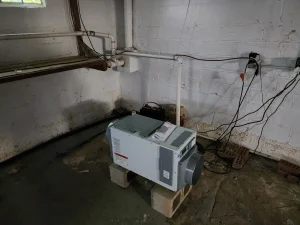 Small dehumidifier on cinder blocks in a dim basement with exposed pipes and a concrete floor. Brick wall shows signs of moisture damage.
