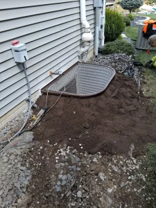 A metal grate covers a basement window beside a house with gray siding. The ground is being dug, and a worker in bright clothing is visible nearby.