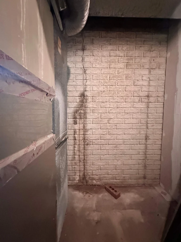 A narrow, dusty space with a brick wall showing water stains. An air duct runs overhead. A single red brick lies on the concrete floor, suggesting neglect.