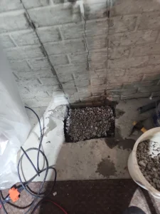 A cracked brick wall with a small hole in the floor filled with gravel. Nearby are cables, a tool, and a bucket of more gravel, suggesting ongoing repairs.