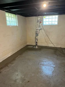 A dimly lit, freshly waterproofed basement with concrete walls and floor, featuring a sump pump in the corner and two glass block windows.