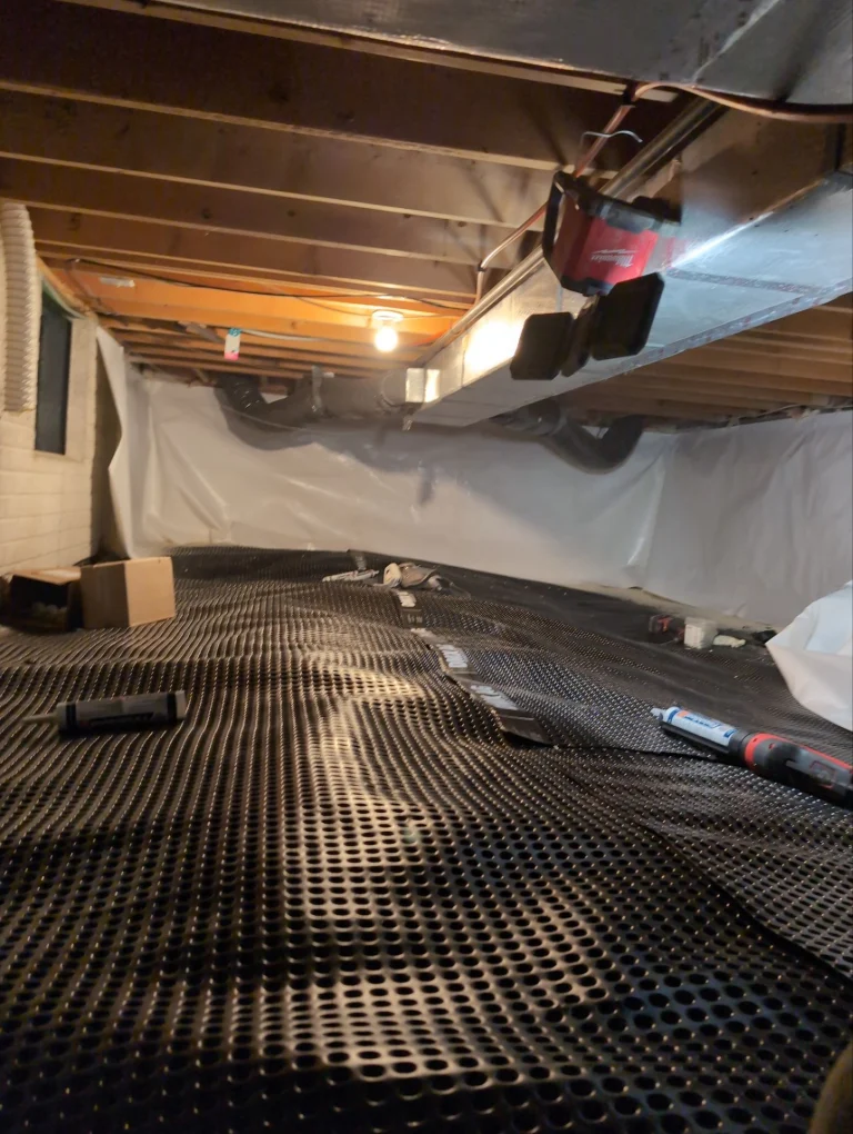 Dimly lit basement with exposed wood beams, ductwork, and a single light bulb. Black, perforated mat covers the floor, with tools scattered around.