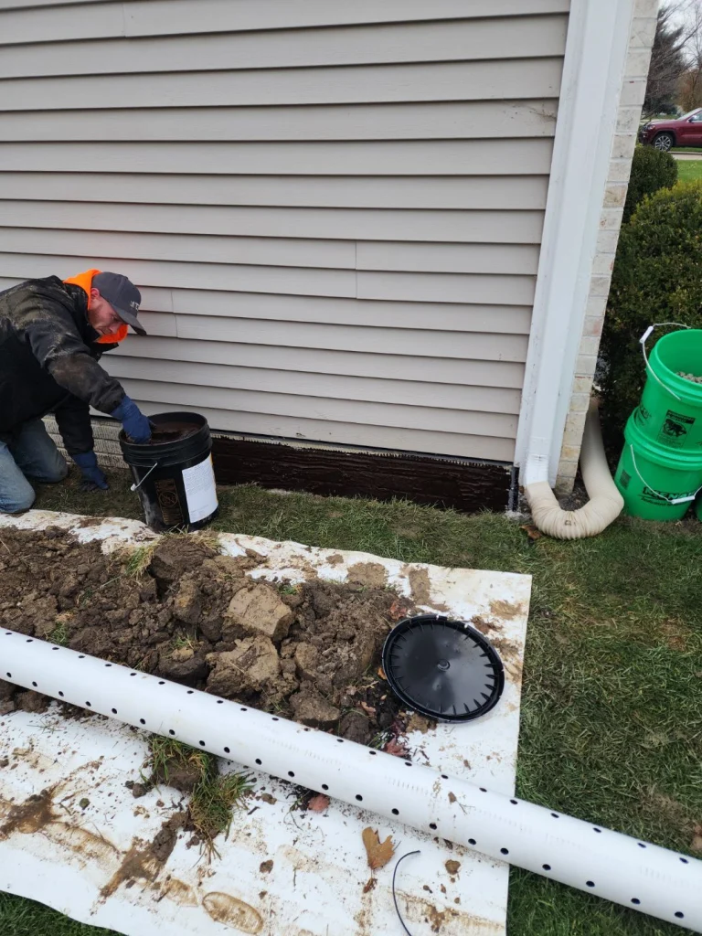 A worker applies a waterproof sealant to a house's foundation near a downspout. Excavated soil and a drainage pipe are on a white tarp nearby.
