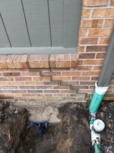 Excavated area near brick foundation showing downspout connection, drainage components, and soil, highlighting foundation repair and drainage improvement efforts.