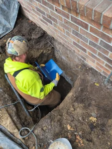Worker excavating near a brick foundation, preparing for helical pier installation to address settling issues at a Perrysburg home.